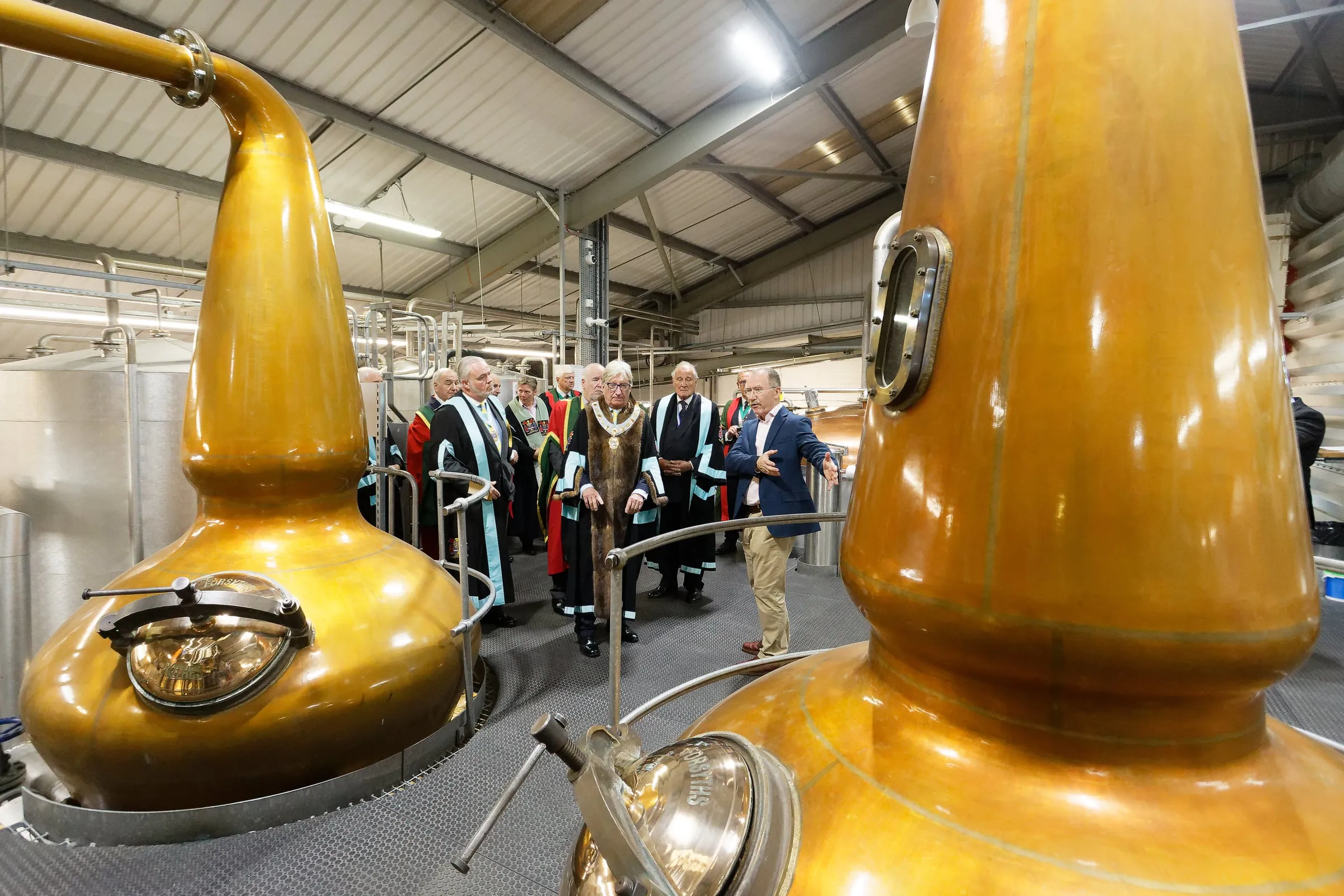 Liverymen in ceremonial gowns visiting a working distillery beside copper pot stills.