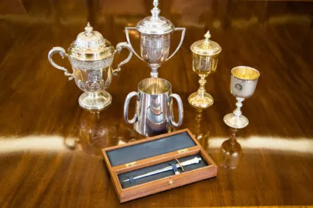 Silver loving cups and other Company plate on a polished table, with a ceremonial dirk in an open case.
