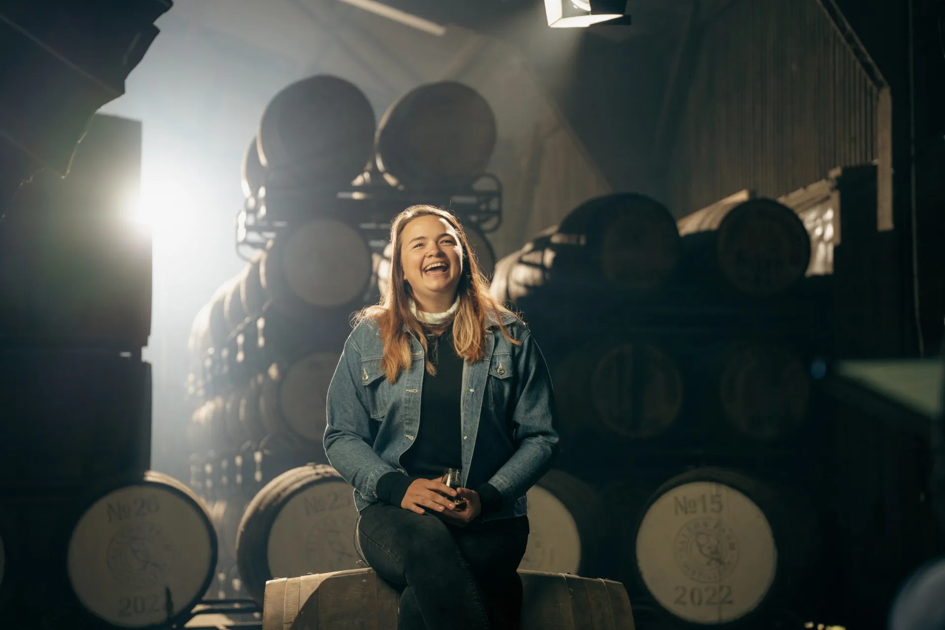 A young woman laughing while seated on a cask in the warehouse at Ellers Farm Distillery.
